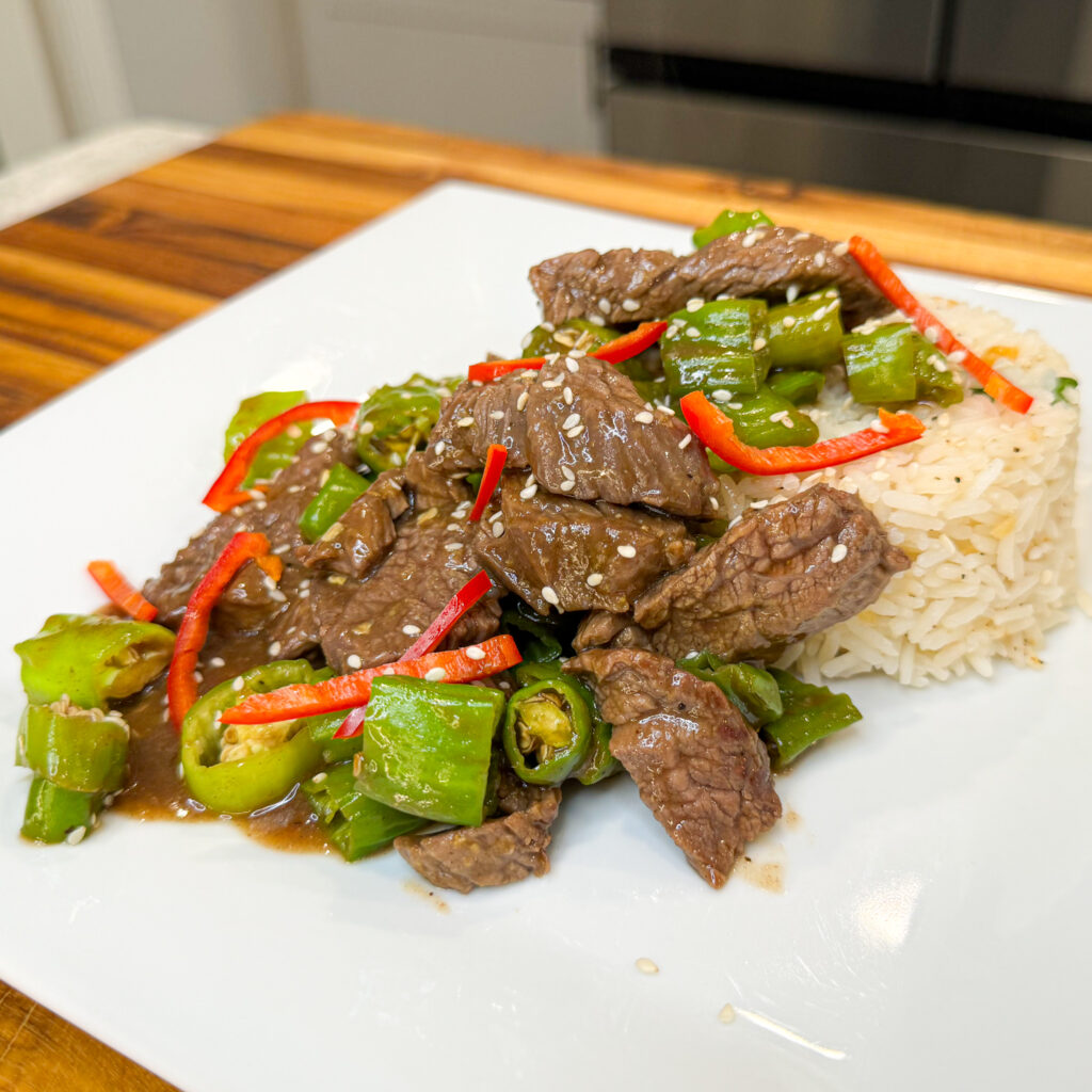 A delicious plate of asian-inspired skirt steak and Shishito peppers featuring tender steak strips seared with shishito peppers, served over a savory rice mixed with peanuts and green onions, and topped with a flavorful ginger-soy sauce and a spicy kick from Serrano peppers.